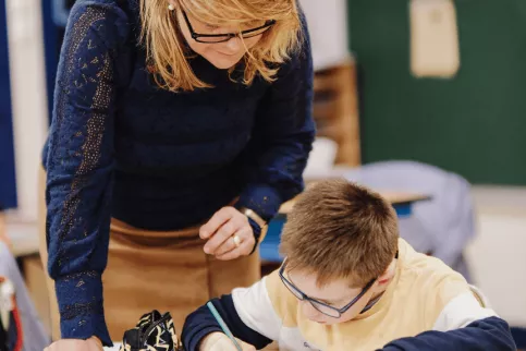 Une AESH accompagne un élève lors des devoirs. Il écrit sur son cahier.